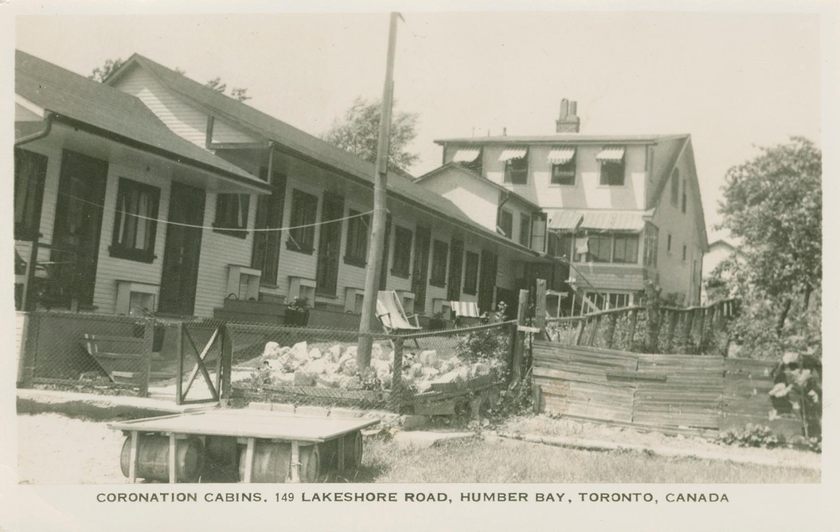 Black and white photograph of a row of summer cottages in front of a larger house.