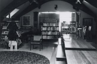 Photo of large room with tales, book shelves and large window with hoarding in background. 