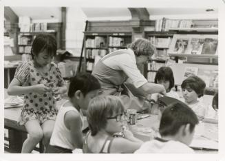 Photo of children seated at a table with library staff member making puppets. 