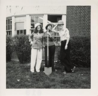 Photo of three women standing on lawn with shovel. 