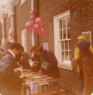 Photo of people browsing a table of books outside library and balloons in background. 