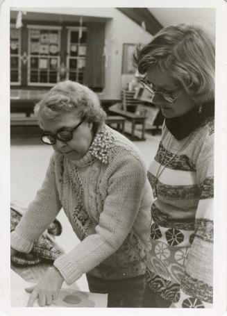 Photo of two women standing looking at a book on a table. 