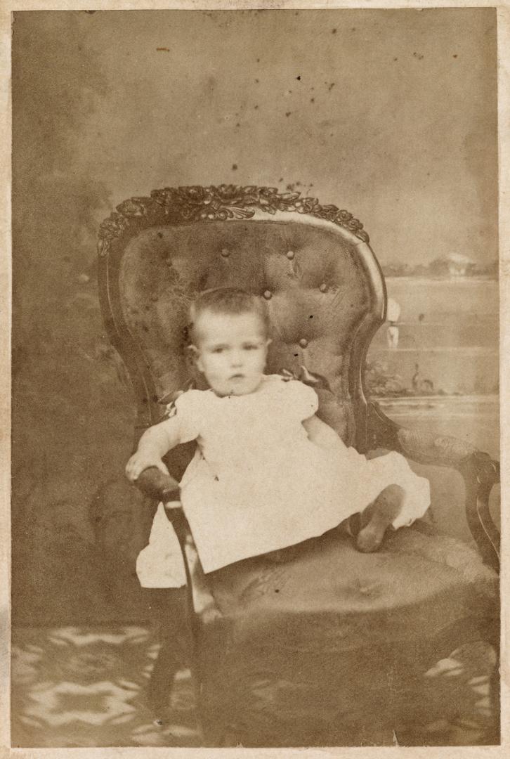 A photograph of an infant wearing a dress and sitting in a cushioned chair in a photography stu…