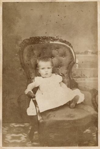 A photograph of an infant wearing a dress and sitting in a cushioned chair in a photography stu ...