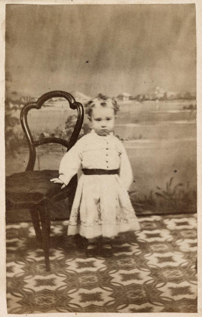 A photograph of a small child standing on a patterned rug, in front of a photo studio backdrop …