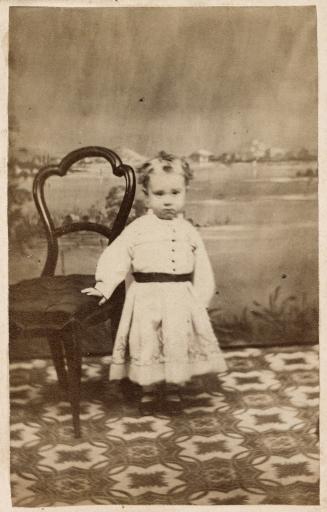 A photograph of a small child standing on a patterned rug, in front of a photo studio backdrop …