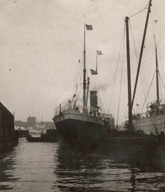 A photograph of a steamship docked in a harbour, with other ships visible to its right and in t ...