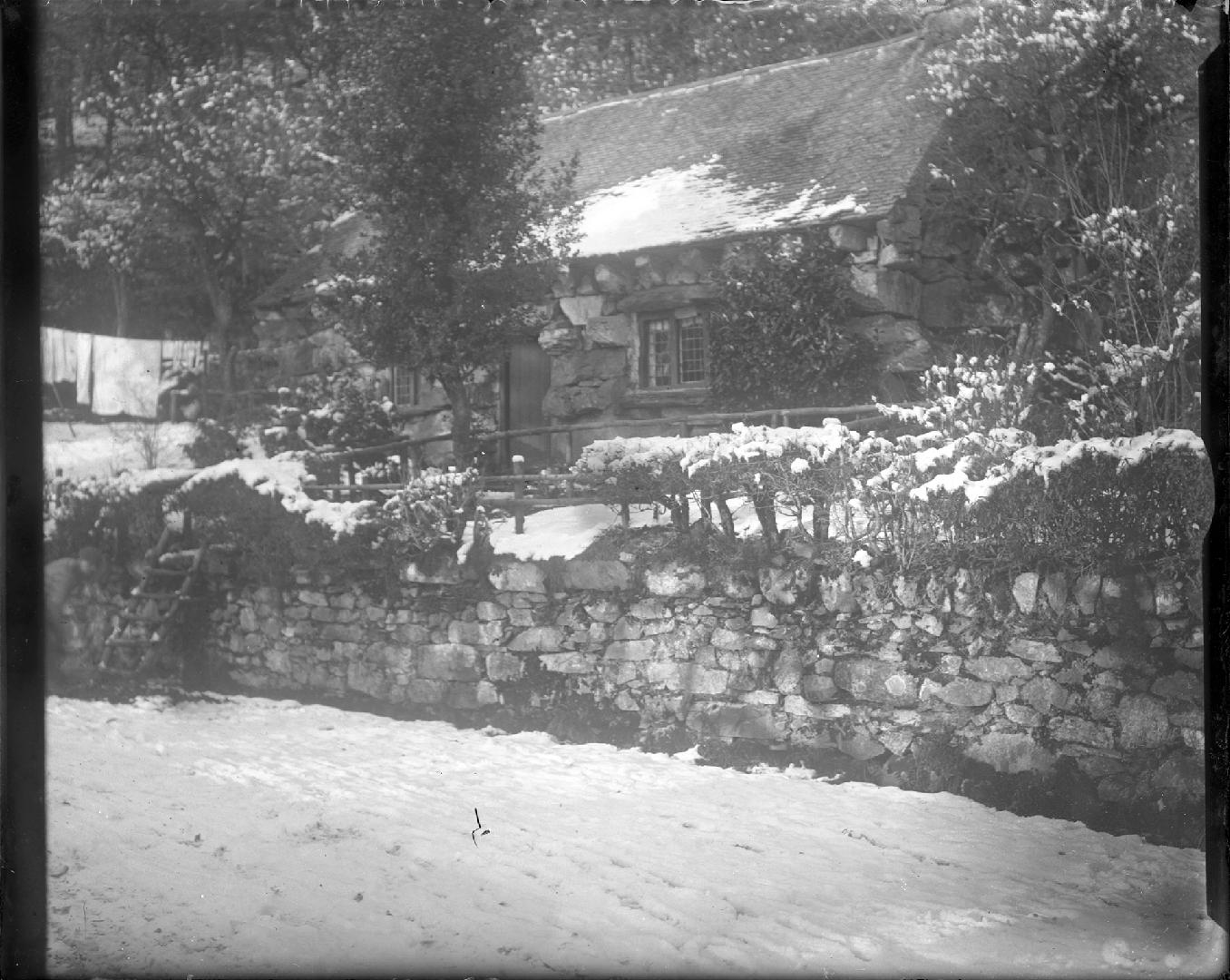 A photograph of a cottage located behind a stone fence approximately four feet tall and some tr ...