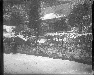 A photograph of a cottage located behind a stone fence approximately four feet tall and some tr ...