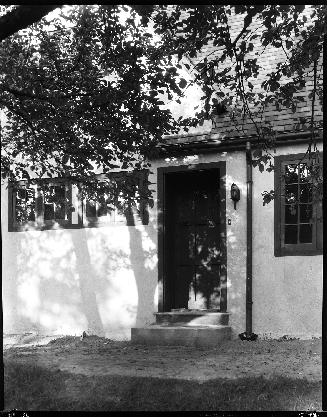A photograph of a door of a house. There are windows on both sides of the door and tree branche ...
