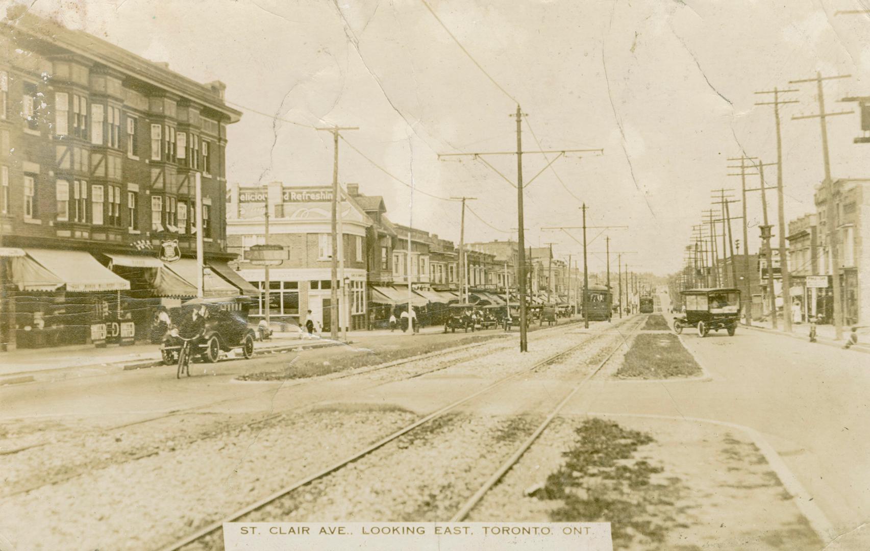 Black and white photograph of a city street with buildings on each side of it, automobiles in t…