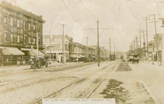 Black and white photograph of a city street with buildings on each side of it, automobiles in t…