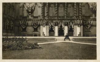 A photograph of the entrance of a large hall, with a lawn in front of it and a garden on the le ...