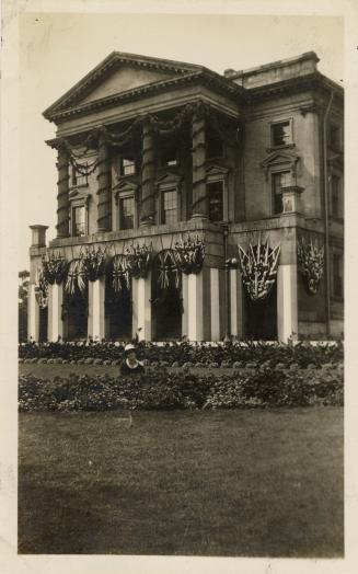 A photograph of a large hall, with a lawn and garden in front of the building. There is a perso ...