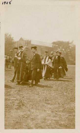 A photograph of a group of people wearing academic robes and mortarboards walking across a gras ...