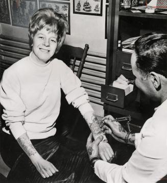 Photograph of a seated woman receiving a tattoo (black and white).