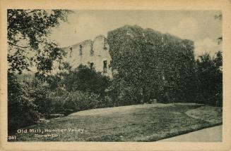 Black and white photograph of a large stone building covered in vines set in a wooded area.