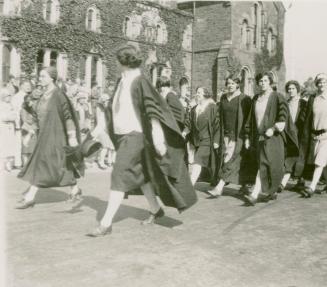 A photograph of a parade through a university campus, with people in academic robes and ties wa ...