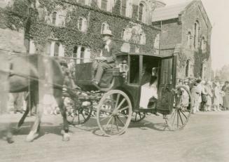 A photograph of a parade through a university campus, with a carriage being pulled by two horse ...