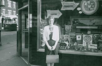 A photograph of a person standing on a city sidewalk in front of a shop window located at the c ...