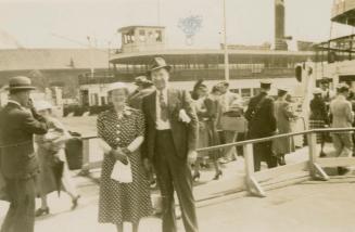 A photograph of two people posing for a photograph in front of two ferries docked in a busy har ...