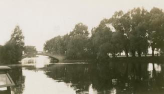 A photograph of a pedestrian bridge spanning a body of water between two islands, with trees an ...