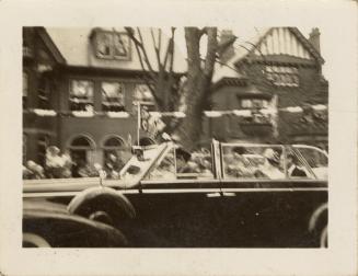 A photograph of a car driving down a city street, with a tree and houses in the background, as  ...