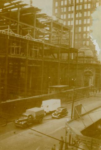 A photograph of a large building under construction, with metal beams exposed. There is a paved ...