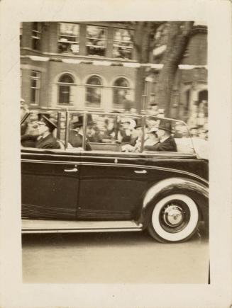 A photograph of a car driving down a city street, with large crowds of people lining the sidewa ...