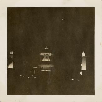A photograph of a large round fountain with two basins, taken at night. The fountain is lit fro ...