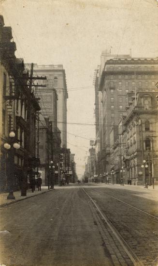 A photograph of a paved city street, with tall buildings on both sides and streetcar tracks in  ...