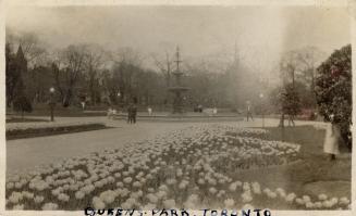 A photograph of a large public park, with flower gardens, paved pathways and a multi-tired foun ...