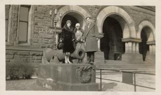 A photograph of three adults standing next to a cannon located next to arched entranceways to a ...