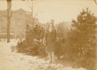 A photograph of a person standing in front of some shrugs and short pine trees. There is a larg ...