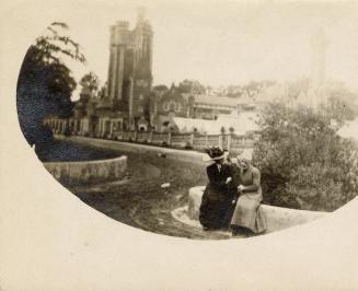 A photograph of two people sitting on a ledge next to a curving path or road leading towards a  ...
