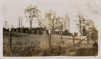 A photograph of a stone castle-like building, with a wire fence, trees and grassy hill in the f ...