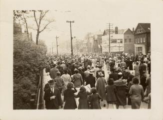A photograph of a large crowd of people walking on and next to a paved city street, with houses ...