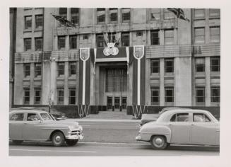A photograph of the bottom three stories of a large building facing a city street, with cars pa ...