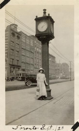 A photograph of a person posing in front of a clock on a tower approximately twenty feet tall,  ...