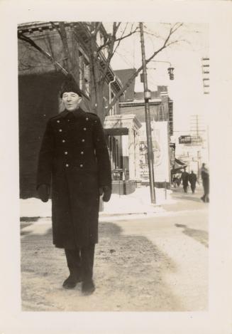 A photograph of a police officer in uniform, standing on a city sidewalk during winter. There i ...
