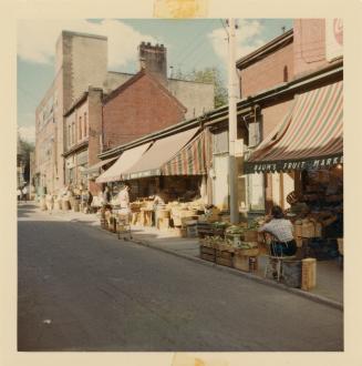 A photograph of a city street, with two and three story buildings on the right side of the road…