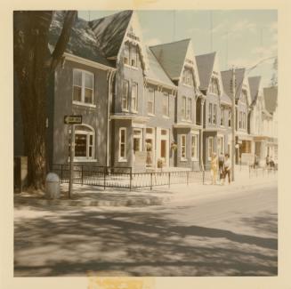 A photograph of a paved city street, with a row of houses on the right side of the street. Ther ...
