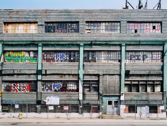 A photograph of a three story warehouse behind a chain link fence, with a sign on the fence ind ...