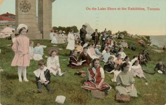 Colorized photograph a group of children in their Sunday best sitting on the grass beside a lak ...