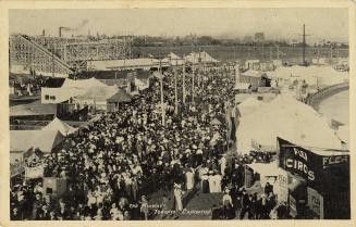 Black and white photograph of crowds of people waling in front of tents and a roller coaster.