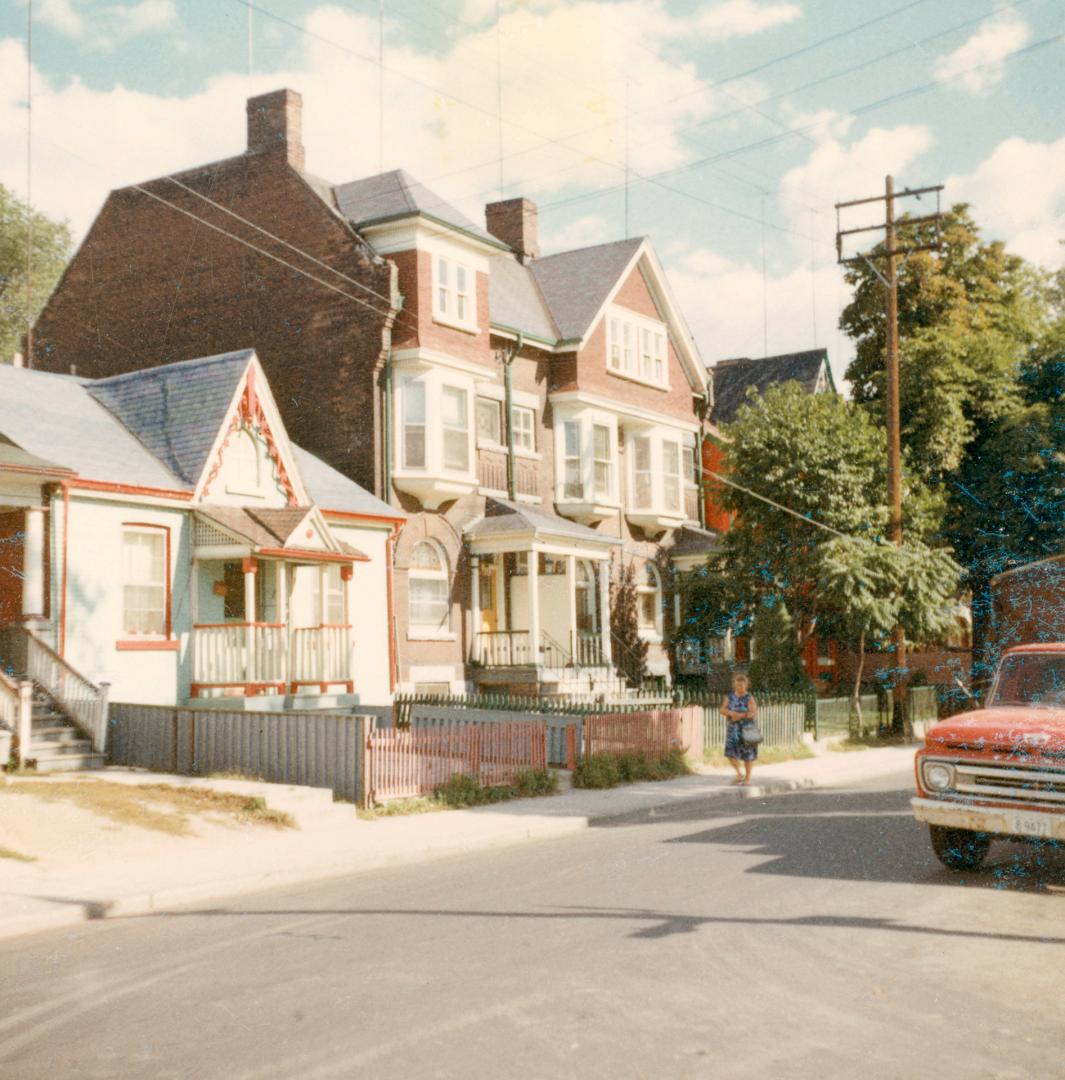 A photograph of a row of two and three story residential houses beside a paved city street. The…