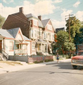 A photograph of a row of two and three story residential houses beside a paved city street. The…