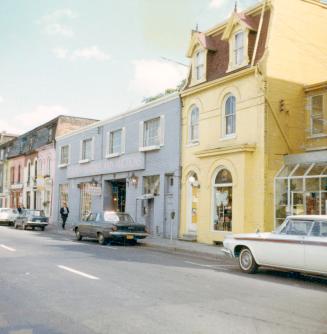 A photograph of a paved city street with a row of two and three story buildings on the far side ...