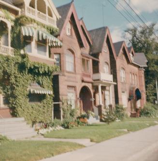 A photograph of a row of three story residential houses, with grass lawns, and a sidewalk in fr ...