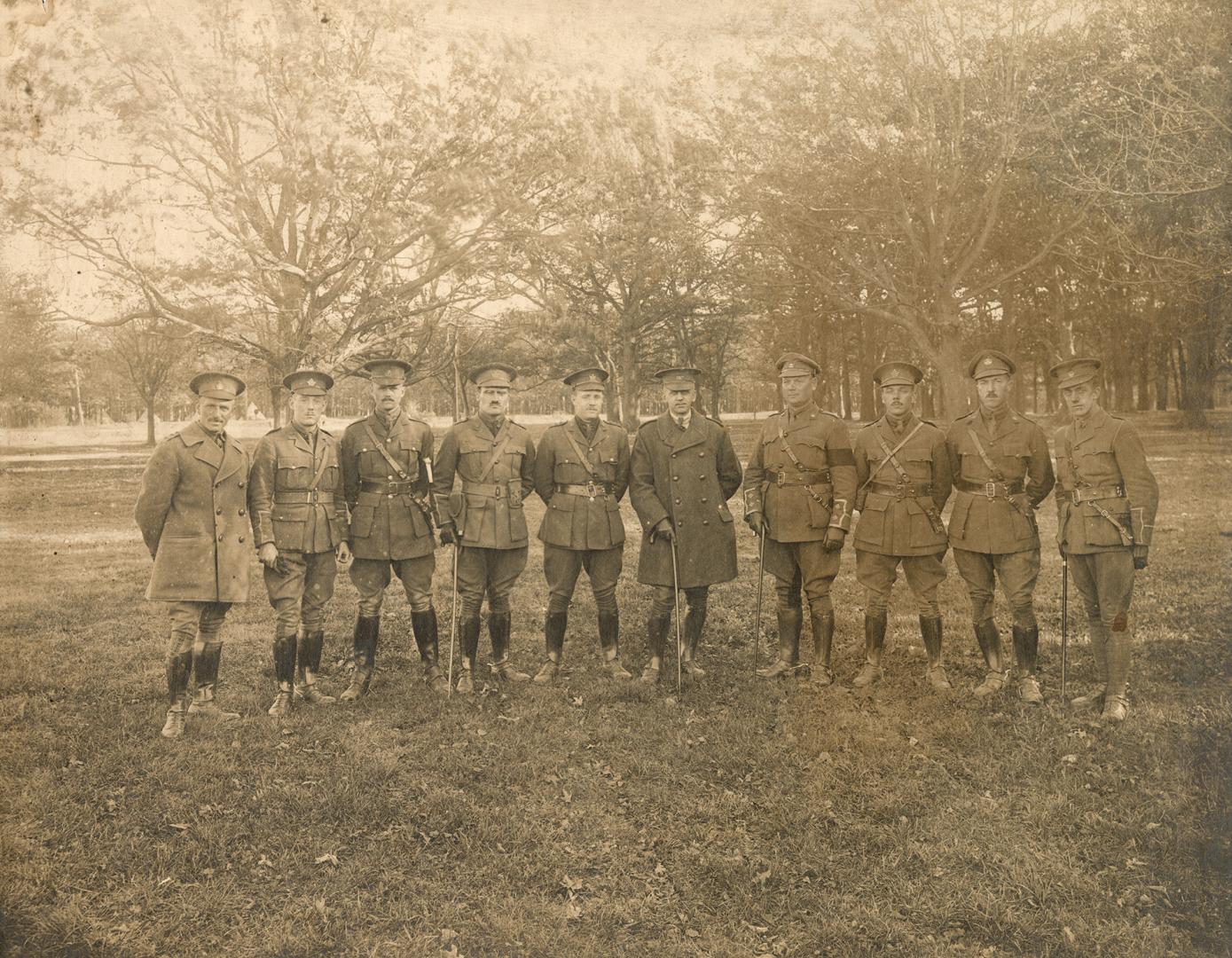 A photograph of ten men posting for a photograph in a grass field surrounded by trees. They are…